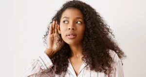Mulher jovem com a mão no ouvido tentando escutar, ilustrando mitos sobre aparelhos auditivos.