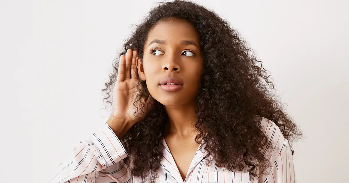 Mulher jovem com a mão no ouvido tentando escutar, ilustrando mitos sobre aparelhos auditivos.
