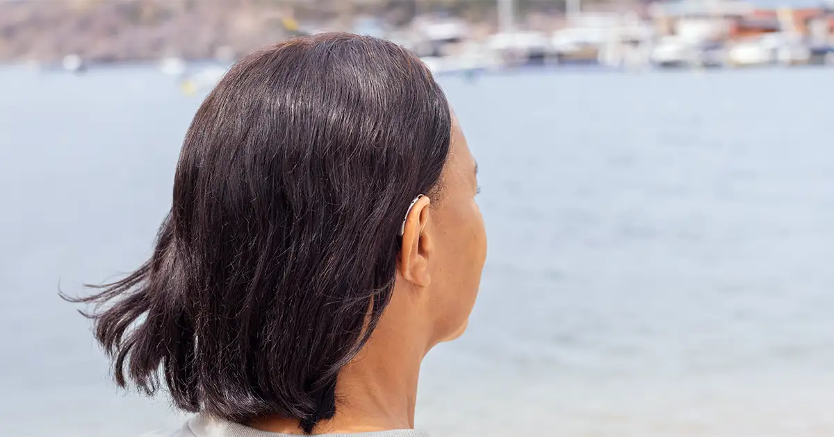 Mulher de costas olhando de forma contemplativa para a água, usando um aparelho auditivo discreto atrás da orelha.