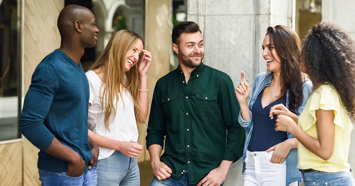 Grupo de cinco jovens adultos sorrindo e conversando animadamente em um ambiente externo.