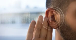 Homem com a mão atrás da orelha tentando ouvir melhor, com gráficos de ondas sonoras ao lado, representando os primeiros sintomas da perda auditiva.